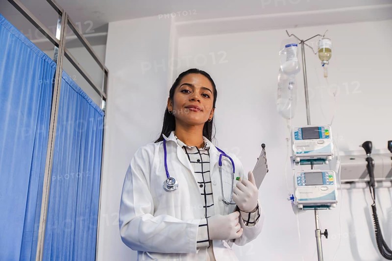 A confident female doctor stands in a hospital setting, holding a clipboard and wearing a white coat and stethoscope, signifying professionalism and healthcare dedication