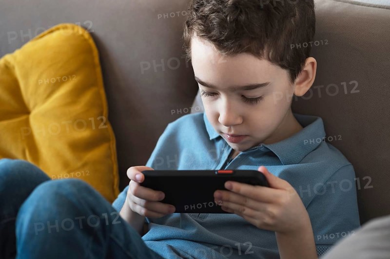 A young boy wearing a blue shirt focuses intently on a smartphone while seated on a comfortable sofa with a yellow cushion, highlighting modern childhood and technology use