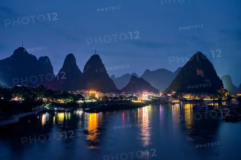 View of Yangshuo town illuminated in the evening with dramatic karst mountain landscape in background over Li river. Yangshuo, China