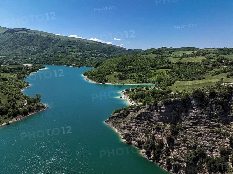A serene capture of Lago di Fiastra in Italy. Vibrant blue waters are surrounded by lush green hills, under a clear blue sky, creating a peaceful, scenic vista