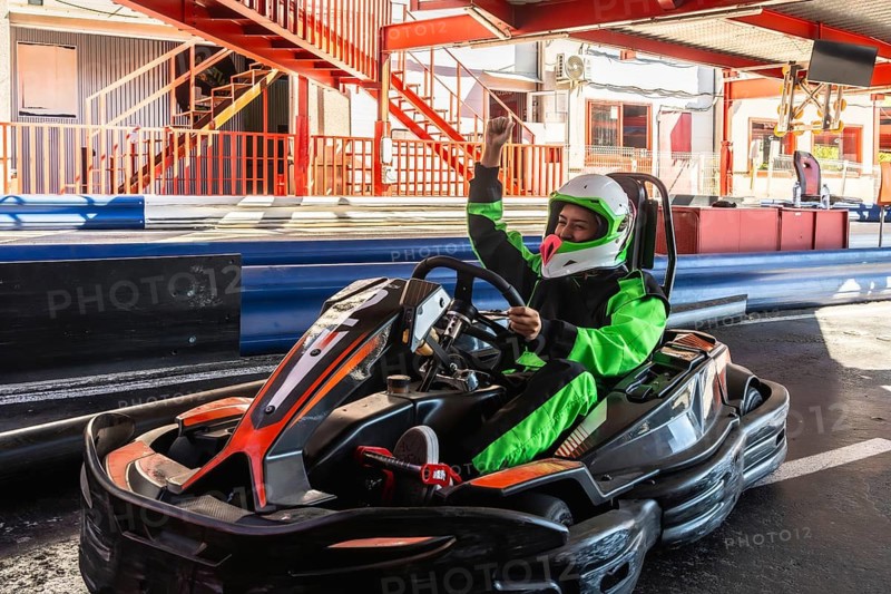 Woman kart driver wearing a helmet and green racing suit raising arm in triumph after winning a race on an outdoor go karting circuit, expressing excitement and success