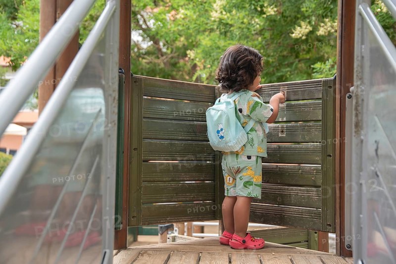 Back view of a young child in colorful attire and backpack explores a wooden playground bridge. Surrounded by lush greenery, the scene captures a moment of curiosity and adventure