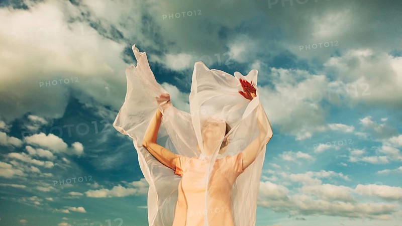 A woman stands with arms raised, her face partially obscured by translucent fabric flowing in the wind. The background features a stunning, cloudy blue sky