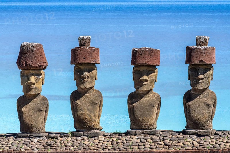 Four Moai statues with red Pukao headpieces on a stone platform in front of a turquoise Pacific Ocean, Ahu Nau Nau, Anakena Beach, Easter Island, Chile