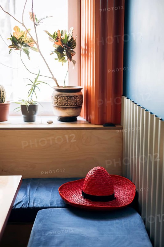 A cozy Mexican interior featuring a vibrant red sombrero on a blue cushioned bench. Sunlight filters through a window with potted plants, creating a warm, inviting atmosphere