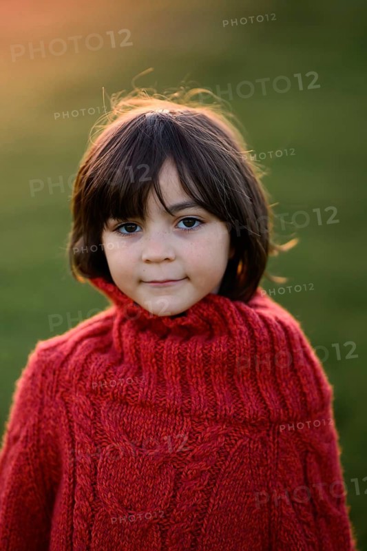 A young girl wearing a vibrant red sweater poses outdoors, her expression thoughtful. The setting sun casts a warm glow, highlighting her dark hair and gentle look. This autumn scene captures the essence of childhood and the beauty of the season