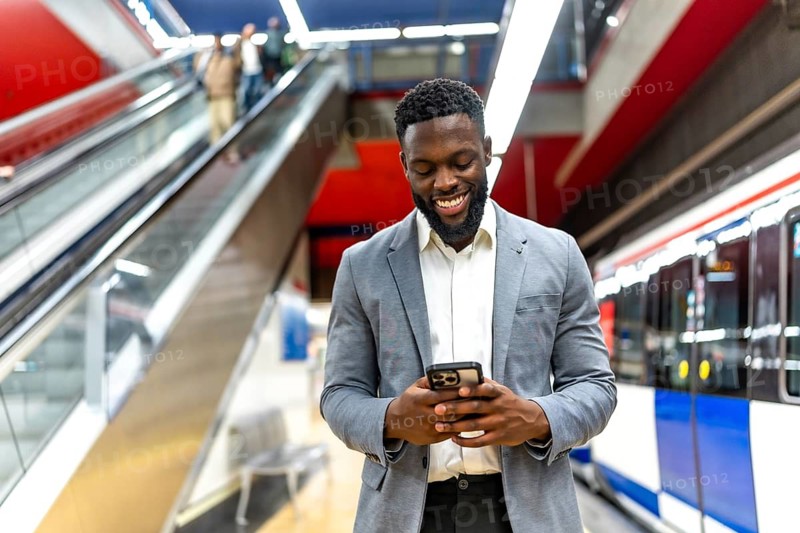Smiling businessman using a smartphone while waiting for a train at an urban subway station, enjoying connectivity and convenience during his commute