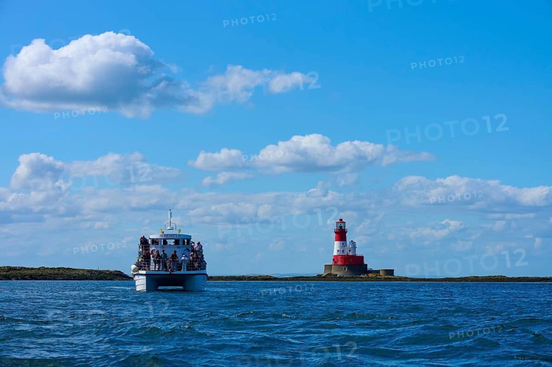 Passenger ship at sea near a red and white lighthouse under blue sky, Seahouses, Longstone Lighthouse, Longstone Rock, Ferns Islands, Northumberland, England, Great Britain