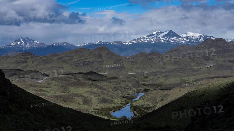 Hilly landscape with a small lake in the valley, snow-capped peaks of the Andes in the background, cloudy sky, Torres del Paine National Park, Patagonia, Chile