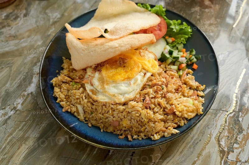 Nasi Goreng Bakso on a blue plate, featuring fried rice with shallots, garlic, sweet soy sauce, and chili. Accompanied vegetables, fried egg, and prawn crackers