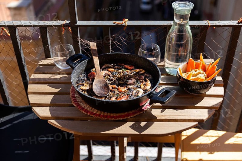 A rustic table on a balcony in Barcelona holds a delicious black paella with seafood dish, water, and appetizers, capturing the essence of Spanish cuisine and outdoor dining enjoyment