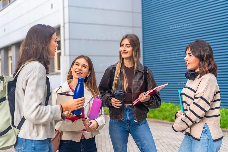 Group of smiling young female university students standing together on campus, carrying backpacks, notebooks, and water bottles while engaging in conversation