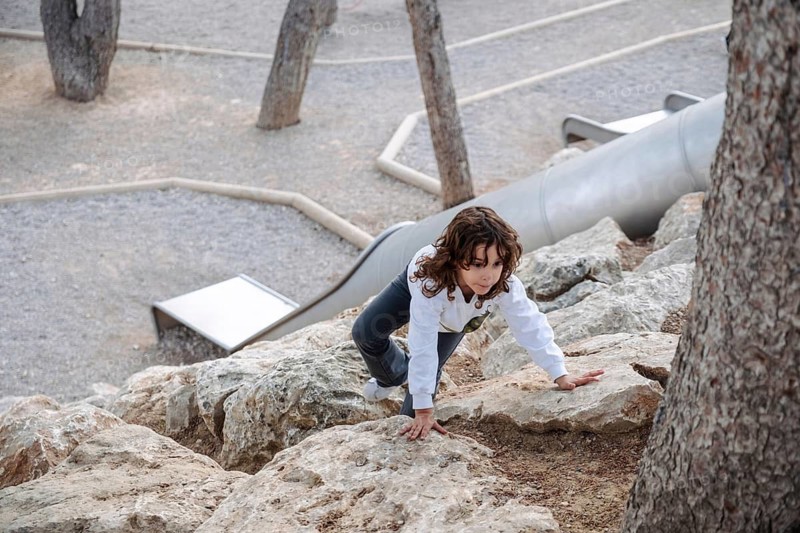 A young girl explores a rocky terrain in a park surrounded by trees. The afternoon light casts a serene ambiance as she navigates the natural environment with curiosity