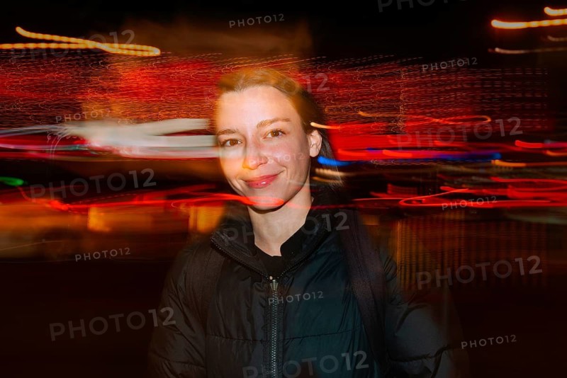 A smiling woman in a black jacket stands against blurred, colorful light trails in an street night setting, creating a vibrant and energetic atmosphere