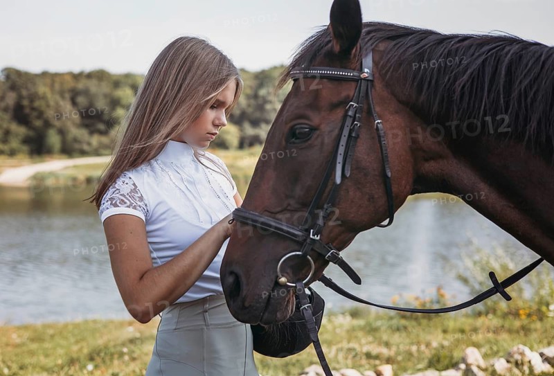 A young girl tenderly interacts with a horse near a tranquil lake. The lush greenery and calm water create a peaceful setting, highlighting the connection between human and animal