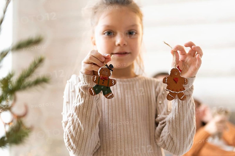 A young girl, looking at the camera, showcases festive gingerbread cookies in a cozy, Christmas-decorated room