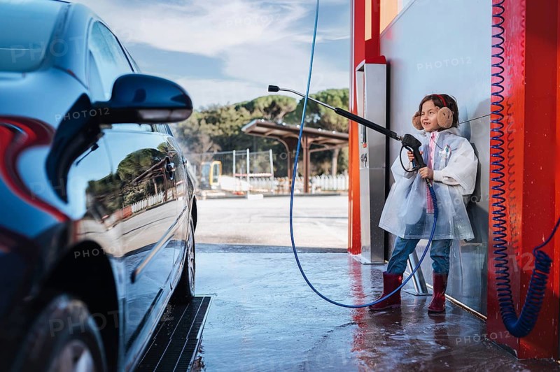 A young girl at a car wash uses a high pressure washer. She smiles, wearing earmuffs and boots, enjoying the task. The reflection of trees is visible on the car