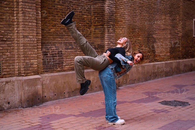 Two young woman friends share a playful moment on a cobblestone street. One joyfully lifts the other, creating a scene filled with laughter and energy against a brick wall backdrop