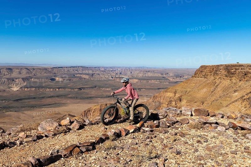 Woman riding a mountain bike, fatbike, on the edge of the abyss, behind the gorge landscape of the Fish River Canyon, Fish River Gorge, Namibia