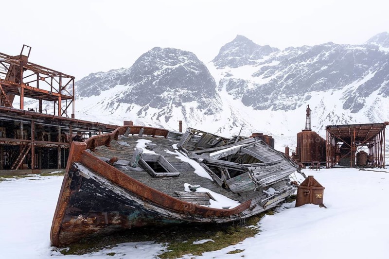 Wreck of a wooden whaling boat in front of rusted facilities of the former whaling station, snow-capped mountains in the background, snowfall, Grytviken, South Georgia