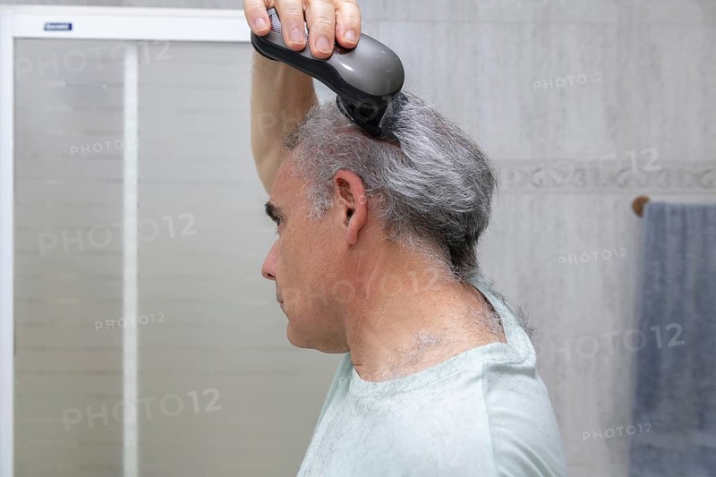 A mature man is seen trimming his hair with an electric trimmer in a tiled bathroom. The image captures the process of self-haircut, highlighting personal grooming