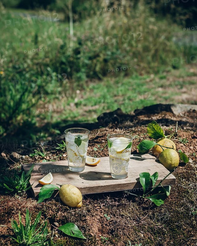 Two glasses of refreshing lemonade with ice and lemon slices sit on a wooden board outdoors. Surrounded by fresh lemons and greenery, it exudes a vibrant, summery feel