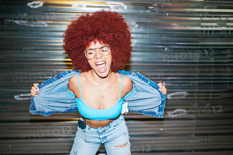 African descendent woman with vibrant afro hair expressing joy, wearing a blue bralette and denim jacket, against a metallic background