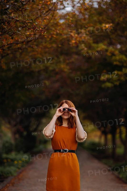 In a serene autumn setting in Quebec, a woman in a vibrant orange dress uses binoculars to explore the colorful foliage. The path is framed by trees with rich fall colors