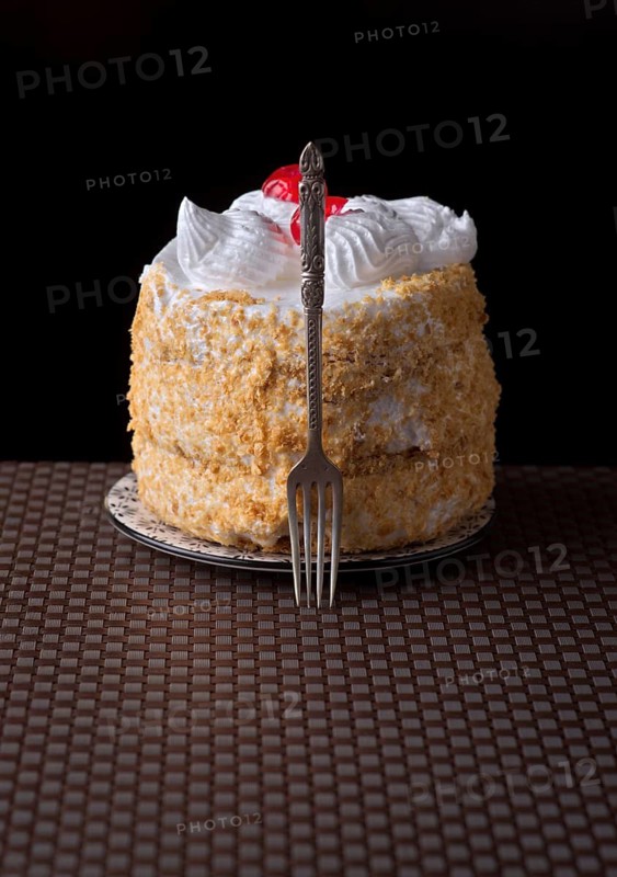 A delectable cake of Tudela, Spain, adorned with meringue and cherries is elegantly showcased with a decorative fork. Set against a dark background, its texture and colors stand out