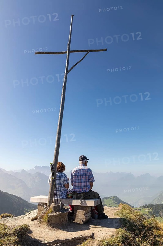 2 hikers sit at the summit cross of the Flügenspitz and enjoy the view, Amden, St. Gallen, Switzerland