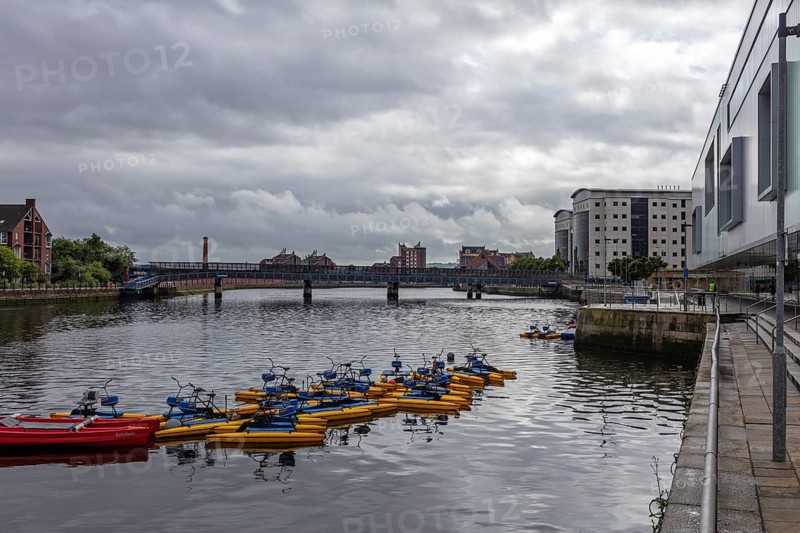 River with anchored boats, surrounded by buildings and a bridge under a cloudy sky, Belfast