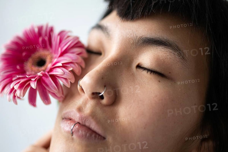 Close-up portrait of an Asian woman with closed eyes. She holds a pink flower near her face, showcasing piercings on her nose and lip, conveying a serene expression