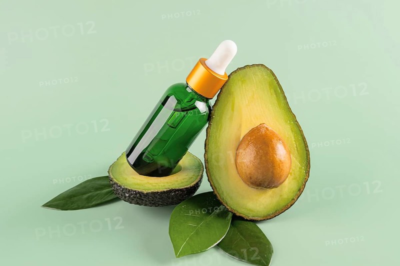 A glass dropper bottle of avocado oil elegantly balanced on half an avocado with green leaves and a soft green backdrop, symbolizing natural skincare and health