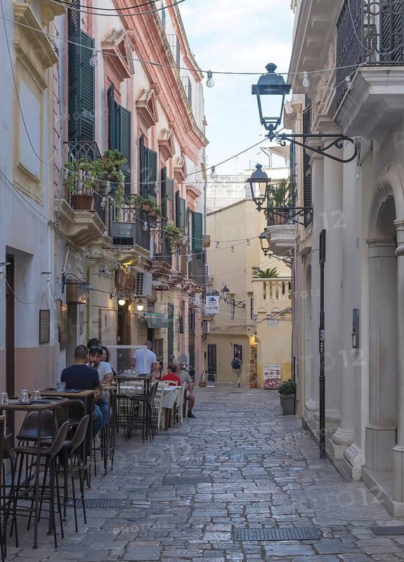 Narrow cobbled alley with cafés and historic Mediterranean-style buildings, Gallipoli, Apulia, Italy