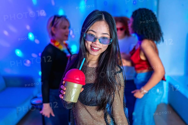 Asian woman holding a colorful slushy drink, smiling and enjoying the vibrant atmosphere of a nightclub party with friends