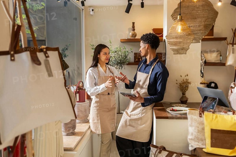 Two employees in aprons converse inside a stylish bag store, surrounded by a variety of bags and warm lighting, creating a welcoming and professional atmosphere