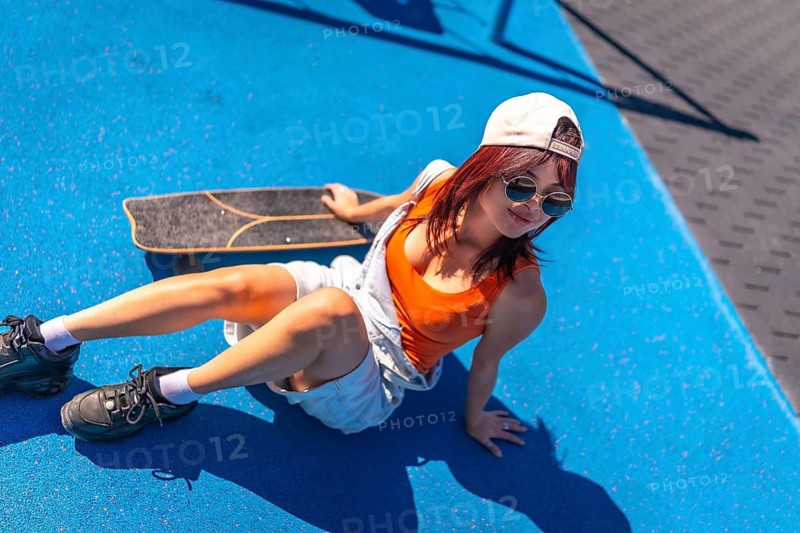 Top view of a distracted young asian woman in denim clohtes sitting next to skateboard in a park