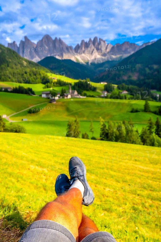 Legs of a tourist relaxing in a meadow with the odle mountains and santa maddalena church in the background, val di funes, dolomites, italy