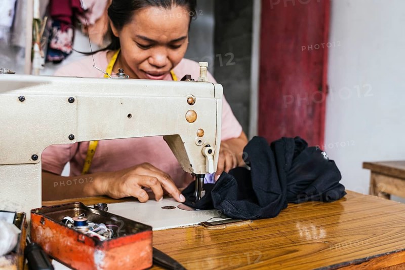 A asian seamstress deeply focused, meticulously sewing fabric with a sewing machine in a vibrant textile workshop, illustrating craftsmanship and dedication in garment making