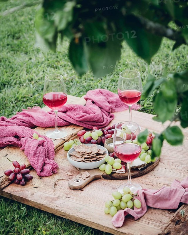 A charming picnic setup featuring glasses of rose, bowls of crispy crackers, and fresh green and red grapes, elegantly arranged on a wooden board, with a lush grass backdrop