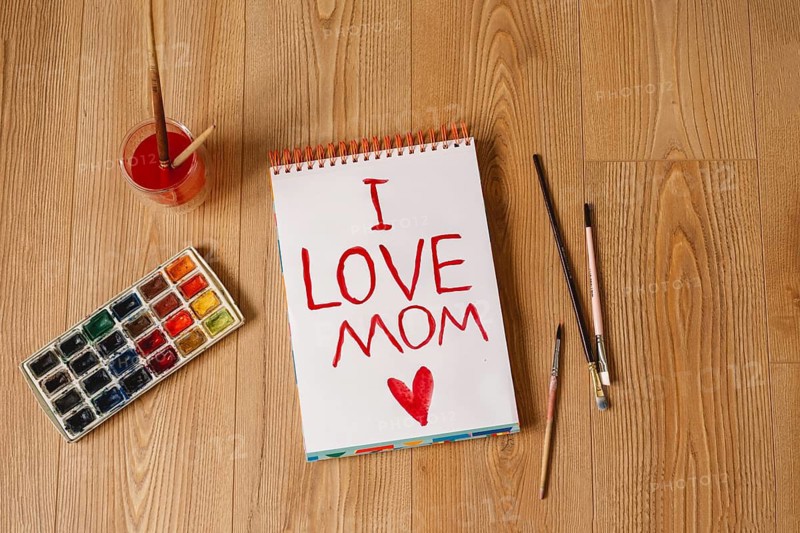 A heartfelt I LOVE MOM message is painted in red on a spiral notebook, surrounded by brushes, watercolor palette, and a paint cup, creating a warm and personal Mother's Day card