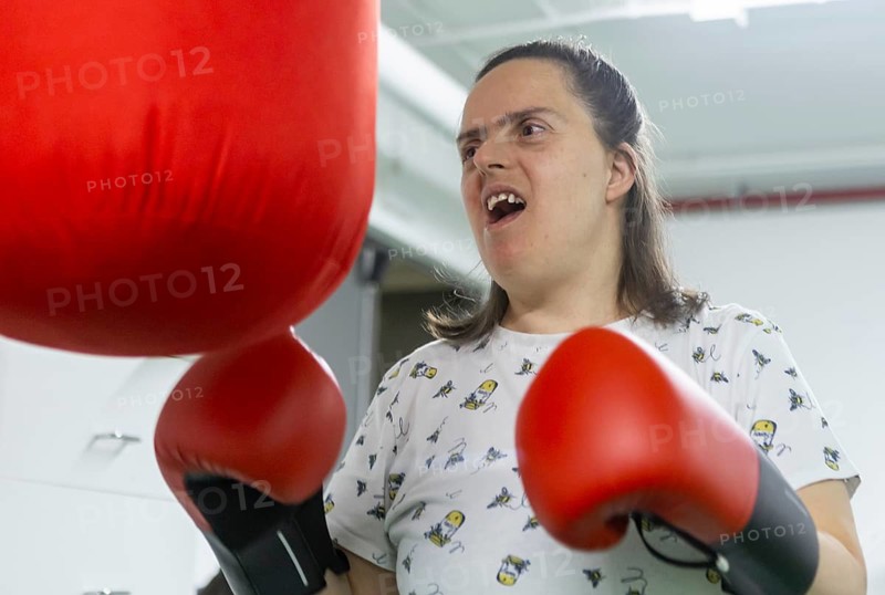 A woman enthusiastically punches a red boxing bag at a mental health residence, using boxing as a form of physical and emotional therapy. They wear a printed T-shirt