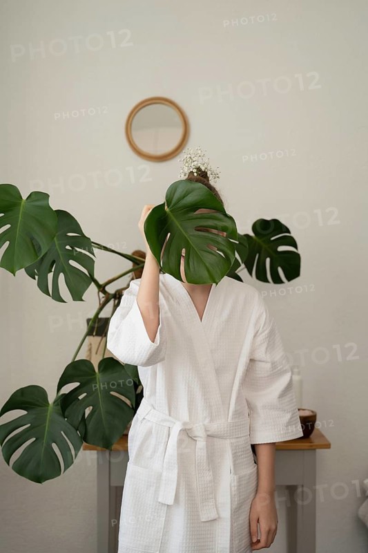A woman in a white bathrobe holds a large monstera leaf covering her face. The setting features a minimalist decor with natural elements and soft light, creating a calm atmosphere