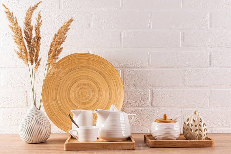 A contemporary kitchen setup displaying ceramic teapots and cups on wooden trays, complemented by dried pampas grass in a textured vase, against a white brick wall background