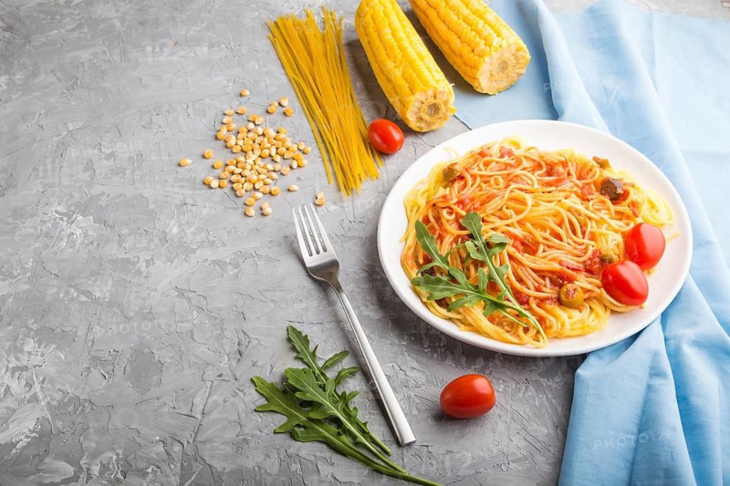 Corn noodles with tomato sauce and arugula on a gray concrete background and blue textile. Side view, copy space