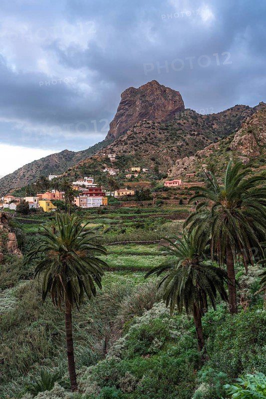 View of Roque El Cano and some houses, palm trees and landscape. Evening, cloudy sky. Vallehermoso, La Gomera, Canary Islands, Spain