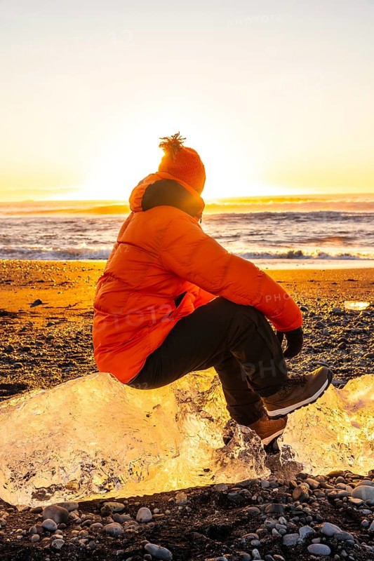 A man in an orange jacket sits on a rock near the ocean. The sun is setting, casting a warm glow over the scene. The man is enjoying the peaceful moment, taking in the beauty of the sunset