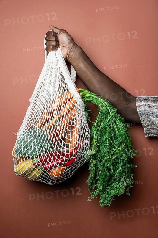 A person holds a reusable mesh bag filled with fresh vegetables, including carrots and peppers, against a brown background. Promotes sustainable shopping and healthy eating
