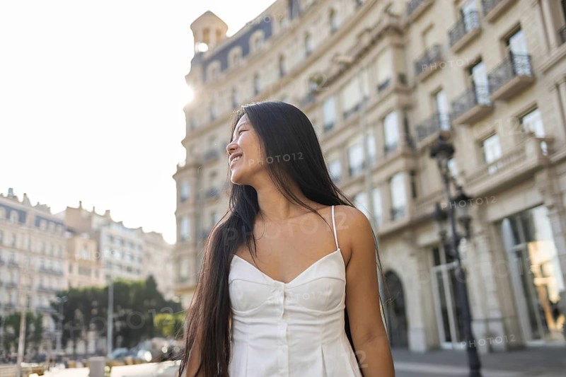 A young Chinese woman with long hair smiles as she enjoys a sunny summer day in the city. Wearing a white dress, she stands against a backdrop of beautiful, historic buildings