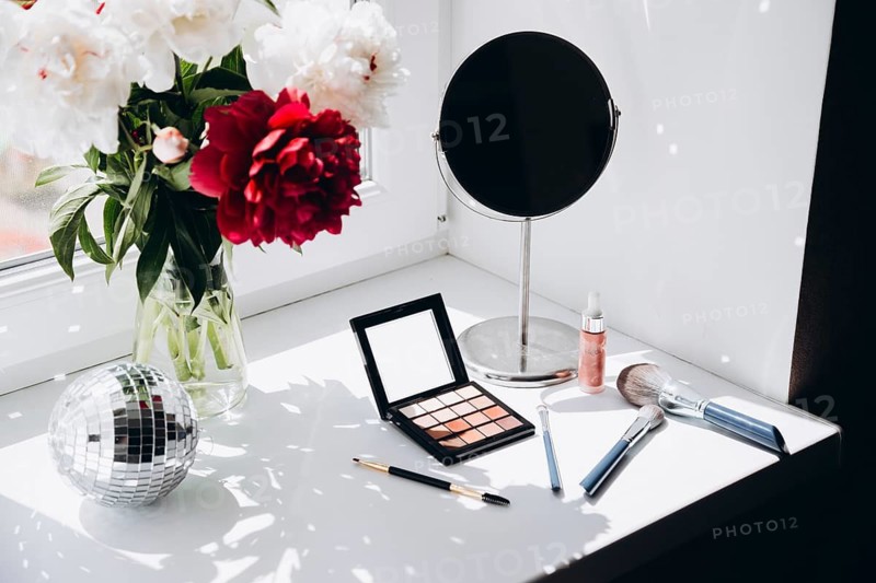 Vibrant red and white peonies in a clear glass vase beside a makeup vanity with a mirror, makeup brushes, and cosmetics, illuminated by natural light from a window. The setup reflects a modern, clean aesthetic ideal for beauty and decor themes